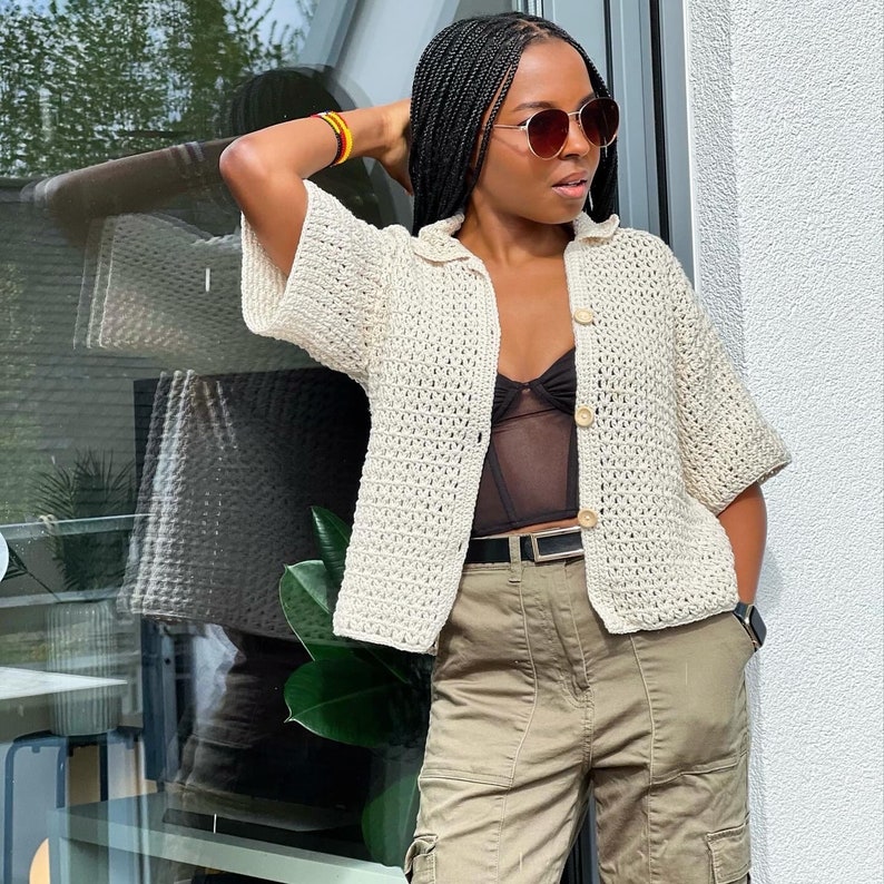 May include: A white crochet cardigan with a button fastening and a black lace bralette underneath. The cardigan has a loose fit and short sleeves. The model is wearing khaki cargo trousers and sunglasses.
