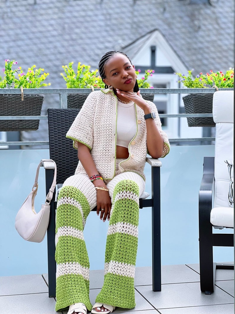 May include: A woman wearing a white and green crochet pantsuit. The pantsuit has a button-up top with short sleeves and wide-leg trousers with horizontal stripes.