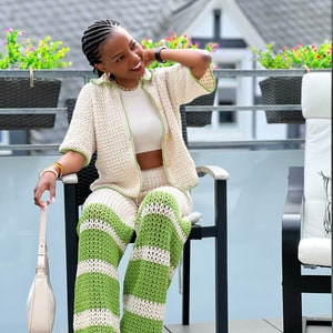 May include: A woman wearing a white crochet top and matching green and white striped crochet trousers. She is sitting on a chair on a balcony.