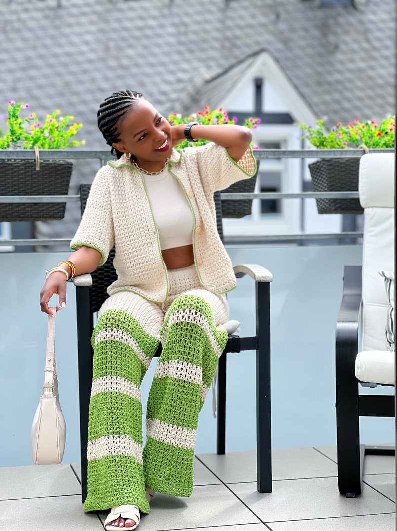 May include: A woman wearing a white crochet top and matching green and white striped crochet trousers. She is sitting on a chair on a balcony.