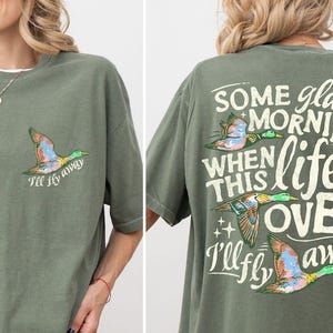 May include: An olive green t-shirt featuring a small flying duck graphic and the text "I'll fly away" on the front left chest. The back displays three flying ducks and the text "SOME glad MORNING WHEN THIS life IS OVER I'll fly away."