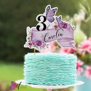 Op de afbeelding: Een verjaardagstaart met lichtblauwe glazuur en een decoratieve topper. De topper heeft een banner met de naam "Carolin", het nummer "3" en paarse vlinders en bloemen. De taart staat op een witte taartstandaard.