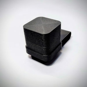 Op de afbeelding: Een zwart, vierkant object met een getextureerd oppervlak en een kleine, rechthoekige uitbreiding. Het object lijkt 3D-geprint te zijn, met zichtbare laaglijnen. Het ontwerp heeft een platte bovenkant en een licht afgeronde basis.