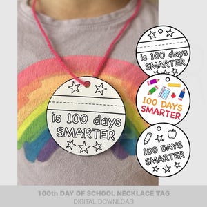 May include: A white, circular necklace tag with the text "is 100 days SMARTER" and star graphics, strung on a pink cord. Additional tags with the same text and school-themed illustrations are also visible. The shirt in the background has a rainbow design.
