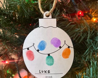 May include: A white circular Christmas ornament with a black string hanger featuring a hand-drawn black string of lights. Four colorful fingerprint light bulbs in red, blue, purple, and orange are present. The text reads LUKE and 2026.