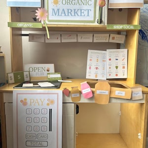 May include: A wooden play market stand with signs that read "Organic Market", "Local Flowers", and "Pay Here". It has shelves with toy food items, a cash register, and a shopping list. The stand is designed for children.