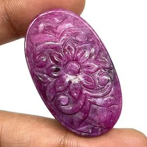Pode incluir: Uma pedra preciosa de formato oval na cor magenta profundo, apresentando um padrão floral detalhado esculpido à mão em sua superfície. A pedra possui um acabamento polido e liso com inclusões naturais visíveis e variações no tom.
