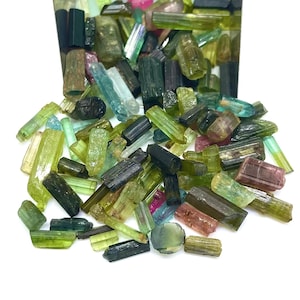 May include: A collection of raw, unpolished tourmaline crystals in various shapes and sizes. The stones display a diverse color palette including shades of forest green, olive, light blue, deep black, and soft pink, with translucent and opaque textures.