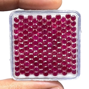 May include: A square clear plastic display case containing a grid of numerous small, round, faceted deep red rubies. Each gemstone is cut with multiple facets to reflect light, arranged in neat rows and columns on a white foam insert base.