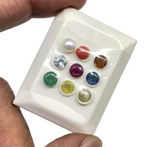 May include: A collection of nine gemstones in a white plastic container. The gemstones include a pearl, a red coral, a clear diamond, a ruby, an emerald, a yellow sapphire, a blue sapphire, a yellow-green sapphire, and a brown gemstone.