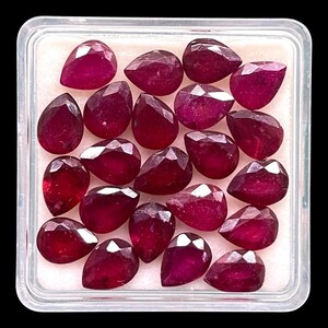 May include: A selection of faceted, teardrop-shaped rubies in a clear plastic container. The gemstones are a deep, rich red colour and appear to be of high quality. The container is square with rounded corners, set against a black background.