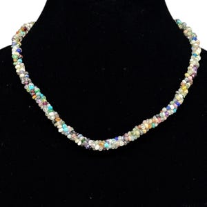 Puede incluir: Un collar de múltiples hilos compuesto por numerosas cuentas pequeñas de piedras preciosas pulidas en una variedad de colores, incluyendo turquesa, azul, morado, amarillo y tonos claros. Las cuentas están dispuestas en un diseño trenzado.
