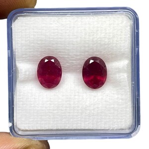 May include: Two oval-cut, deep red rubies are displayed side-by-side within a clear plastic storage box. The gemstones feature a faceted surface that reflects light, and they are set against a white, textured foam insert inside the container.