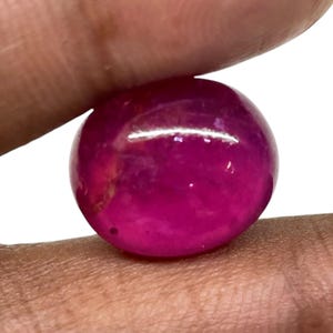 May include: A polished, round, deep magenta gemstone. The gem is held between fingers, showcasing its smooth, reflective surface. The stone's colour is a vibrant, saturated hue, with subtle variations in tone.