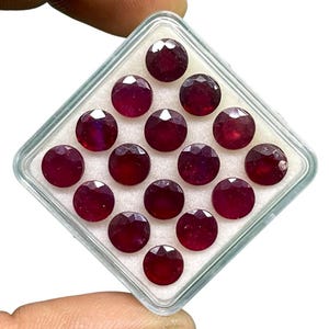 Pode incluir: Uma coleção de quatorze pedras preciosas redondas, facetadas, vermelho-escuras, possivelmente rubis, é exibida em um recipiente de plástico transparente e quadrado com interior branco. As pedras preciosas estão dispostas em um padrão de grade, mostrando suas superfícies polidas.
