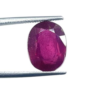 May include: A deep magenta-coloured oval gemstone with a faceted surface held by a pair of silver-toned metal tweezers. The stone displays a translucent quality with visible internal inclusions and sharp, geometric cut facets across its top.