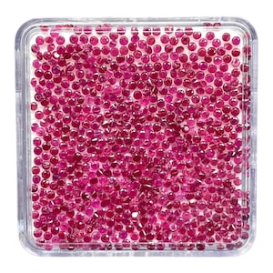 May include: A square clear plastic display case filled with a large collection of small, round, faceted loose rubies. The gemstones exhibit a vibrant, deep pinkish-red colour and are arranged in a dense, uniform layer within the container.
