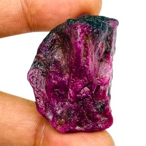 May include: A raw, unpolished gemstone with a deep magenta and dark purple colour palette. The stone features a rough, crystalline texture with natural facets and irregular edges, displaying varying levels of translucency and dark inclusions.