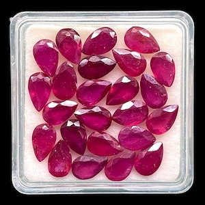 May include: A collection of faceted, pear-shaped rubies in a clear plastic container. The gemstones are a deep, vibrant red color, with multiple facets catching the light. The container sits on a white surface against a black background.