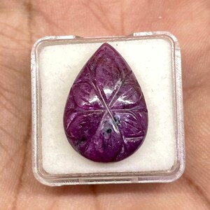 May include: A teardrop-shaped, deep magenta ruby gemstone featuring a carved six-pointed star pattern on its surface. The gemstone is housed within a small, square, clear plastic display box with a white foam insert for secure placement.