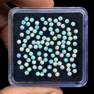 May include: A collection of small, round opals in a clear plastic container. The opals are white with iridescent flashes of blue, pink, and green. The container is square with a black interior, and the opals are densely packed.