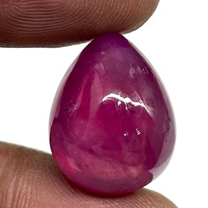 May include: A pear-shaped, cabochon-cut gemstone with a deep, vibrant magenta-pink colour. The stone features a smooth, polished surface with a high-gloss finish that reflects light, showcasing its translucent quality and rich, saturated hue.
