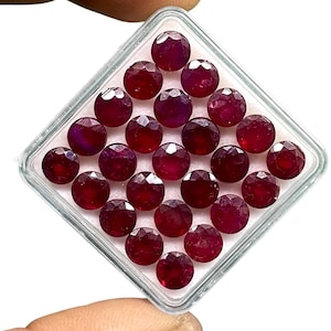 Pode incluir: Uma coleção de pedras preciosas facetadas, redondas, vermelho escuro, exibidas em um recipiente de plástico transparente e quadrado. As gemas têm uma superfície polida e reflexiva, e são dispostas em fileiras organizadas. O recipiente é segurado por uma mão, em um fundo branco.