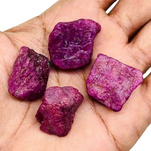 May include: Four raw, unpolished ruby gemstones with a deep magenta-red color and irregular, jagged surfaces. These natural mineral specimens display a coarse, crystalline texture with varying shades of dark pink and purple throughout each piece.