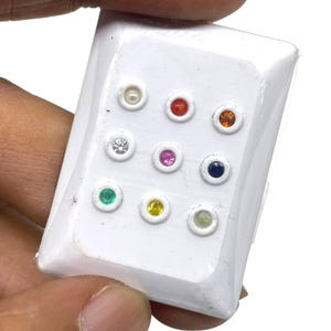 Puede incluir: Una tarjeta de presentación rectangular blanca con nueve piedras preciosas pequeñas, redondas y facetadas dispuestas en una cuadrícula de tres por tres. Las piedras incluyen una perla blanca, y gemas de color rojo, naranja, transparente, rosa, azul oscuro, verde, amarillo y verde claro.