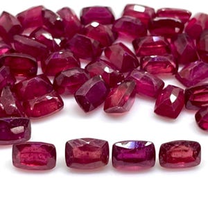 May include: A selection of faceted, deep red ruby gemstones. The gemstones are scattered on a white surface, displaying their various shapes and the light reflecting across their surfaces. The gems have a polished, reflective quality.