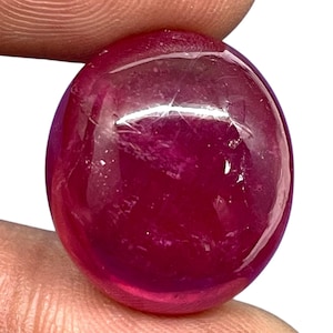 Pode incluir: Uma pedra preciosa redonda lapidada em cabochão com uma cor vermelho-magenta profunda e saturada. A superfície da pedra é lisa e polida, exibindo inclusões naturais internas e reflexos de luz sobre a sua forma arredondada e convexa.