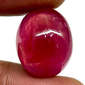 Può includere: Un rubino ovale e lucido. La gemma è di un colore rosso intenso e saturo, con una superficie liscia e lucente. Il rubino è tenuto tra due dita, evidenziando la sua forma e il suo colore.