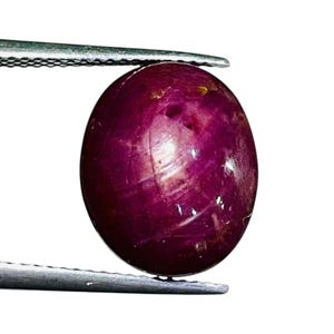Könnte beinhalten: Ein tief magentafarbener, ovaler Cabochon-Edelstein mit einer glatten, polierten Oberfläche und sichtbaren Einschlüssen. Der Stein wird zwischen den Metallspitzen einer Pinzette gehalten und zeigt seinen satten, rot-violetten Farbton.