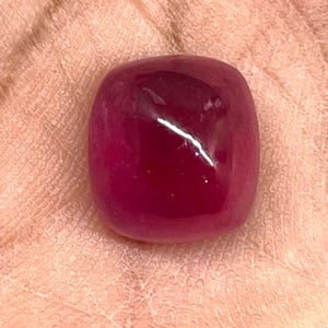 May include: A deep magenta-coloured gemstone with a smooth, rounded cushion-cut surface. The stone features a polished, reflective finish that catches the light, displaying a rich, saturated red-pink hue across its entire rounded square shape.