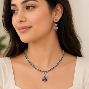 May include: A silver-toned necklace and matching drop earring set featuring alternating round-cut green and pink gemstones. The necklace has a central floral-shaped pendant, while the earring features a small vertical drop with a floral cluster.