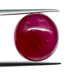 Könnte beinhalten: Ein runder, im Cabochon-Schliff gehaltener Edelstein in einer tiefen, gesättigten roten Farbe. Der Stein hat eine glatte, polierte Oberfläche mit einem subtilen Glanz und wird zwischen den zwei Metallspitzen einer silberfarbenen Pinzette gehalten.