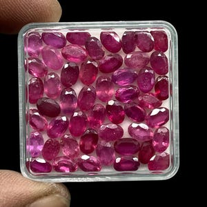 May include: A collection of oval-cut rubies in a clear plastic container. The gemstones are a deep red-pink color and are arranged closely together. The container is held by a person's hand against a black background.