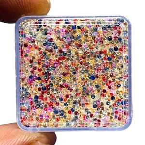 May include: A square, clear plastic container filled with a large collection of small, faceted, round gemstones in a variety of colours including red, blue, yellow, pink, and clear, arranged in a dense, mixed pattern within the box.