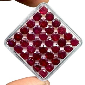May include: A collection of faceted, deep red gemstones, likely rubies, arranged in a clear, square plastic container. The gems are round with multiple facets, reflecting light. The container is held by a hand, with a white background.