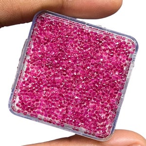 May include: A square, clear plastic container filled with a large quantity of small, round, faceted, vibrant pink gemstones. The stones are densely packed together, displaying a uniform bright pink colour and reflective surfaces throughout the box.