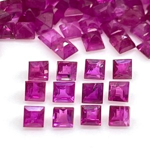 May include: A collection of square-cut, deep magenta rubies. The gemstones are arranged in a scattered pattern, with some individual stones and others grouped together. The facets of the rubies catch the light, creating a sparkling effect. The gems are set against a white background.