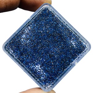 May include: A clear, square container filled with numerous small, dark blue beads. The beads are densely packed, creating a textured surface. The container is held by a person's fingers against a white background.
