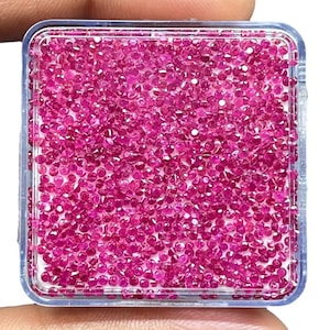 May include: A square, clear plastic container filled with a large collection of small, round, faceted loose rubies. The gemstones are a vibrant, deep pinkish-red colour and are packed closely together, covering the entire base of the storage box.