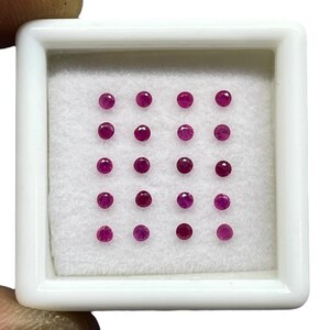 May include: A collection of twenty small, round, faceted rubies arranged in a five-by-four grid inside a square white display box. The gemstones are deep red and set against a white felt-like lining within the white plastic container.