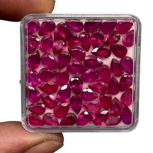 May include: A collection of faceted, pear-shaped rubies in a clear plastic container. The rubies are a deep, rich red color and appear to be of high quality. The container is held by a person's fingers.