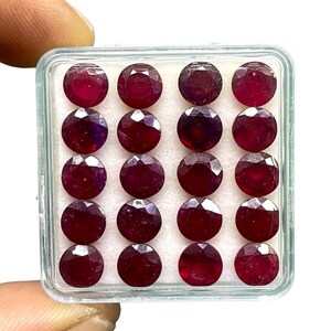 May include: Sixteen round, faceted, deep red gemstones, possibly rubies, are presented in a clear plastic container. The gems have a polished, reflective surface and are arranged in a grid pattern against a white background.