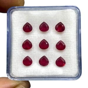 May include: A collection of nine loose, pear-shaped red rubies arranged in a three-by-three grid inside a square, clear plastic display case with a white foam insert. Each gemstone features a faceted surface that reflects light.