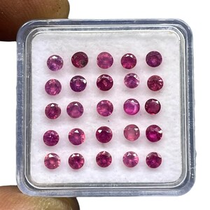 Puede incluir: Una colección de rubíes redondos y facetados en varios tonos de rojo y rosa, exhibidos en una caja de plástico transparente con interior blanco. Las piedras preciosas son de tamaño y forma similares.