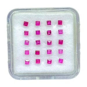 Puede incluir: Una caja de presentación cuadrada de plástico transparente que contiene veinte pequeñas piedras preciosas de color rosa vibrante, con talla princesa, dispuestas en una cuadrícula de cinco por cuatro sobre un inserto de espuma blanca. Cada gema tiene facetas.