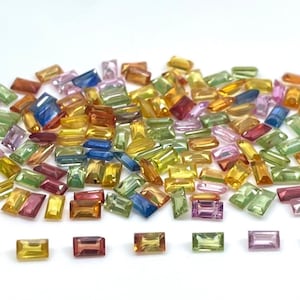 May include: A collection of small, rectangular, faceted gemstones in a variety of colors including yellow, green, blue, pink, and orange. The stones are arranged in a large, loose pile with seven individual stones lined up in a row below.